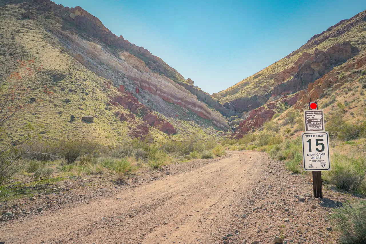 A dirt road running up into Jawbone Canyon between steep, banded red and tan hillsides, past a Friends of Jawbone 'pack it in, pack it out' sign and a 'speed limit 15 near camp areas' sign.