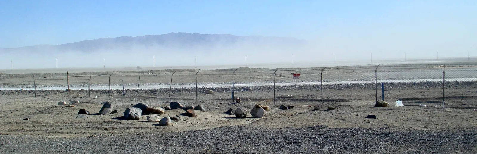 A wall of blowing dust rising off a flat dry lake bed and hiding the foot of a mountain range, seen past a chain-link fence and scattered boulders.