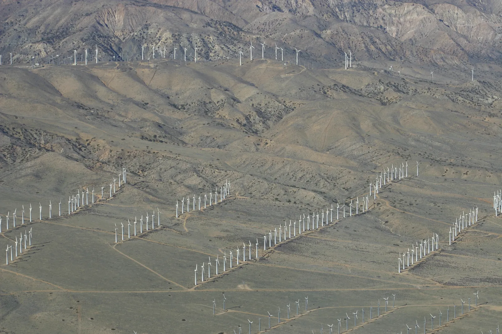 Aerial view of bare, dry foothills crossed by long rows of white wind turbines, with more turbines along the ridge of the mountains behind.