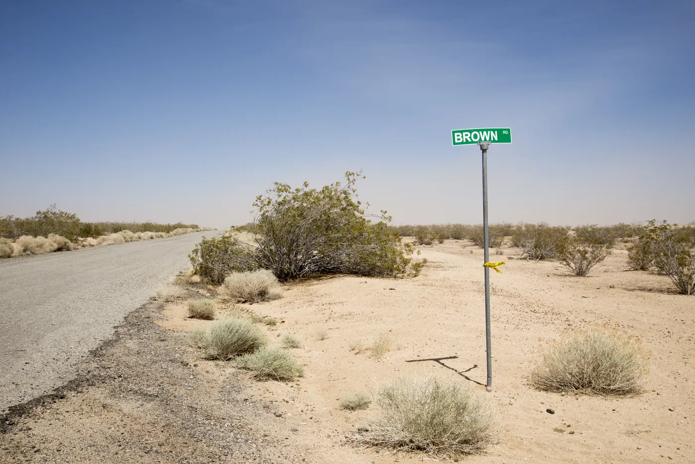 A green street sign reading Brown Rd on a pole in open sand and creosote bushes beside a narrow paved road, with no buildings anywhere in view.