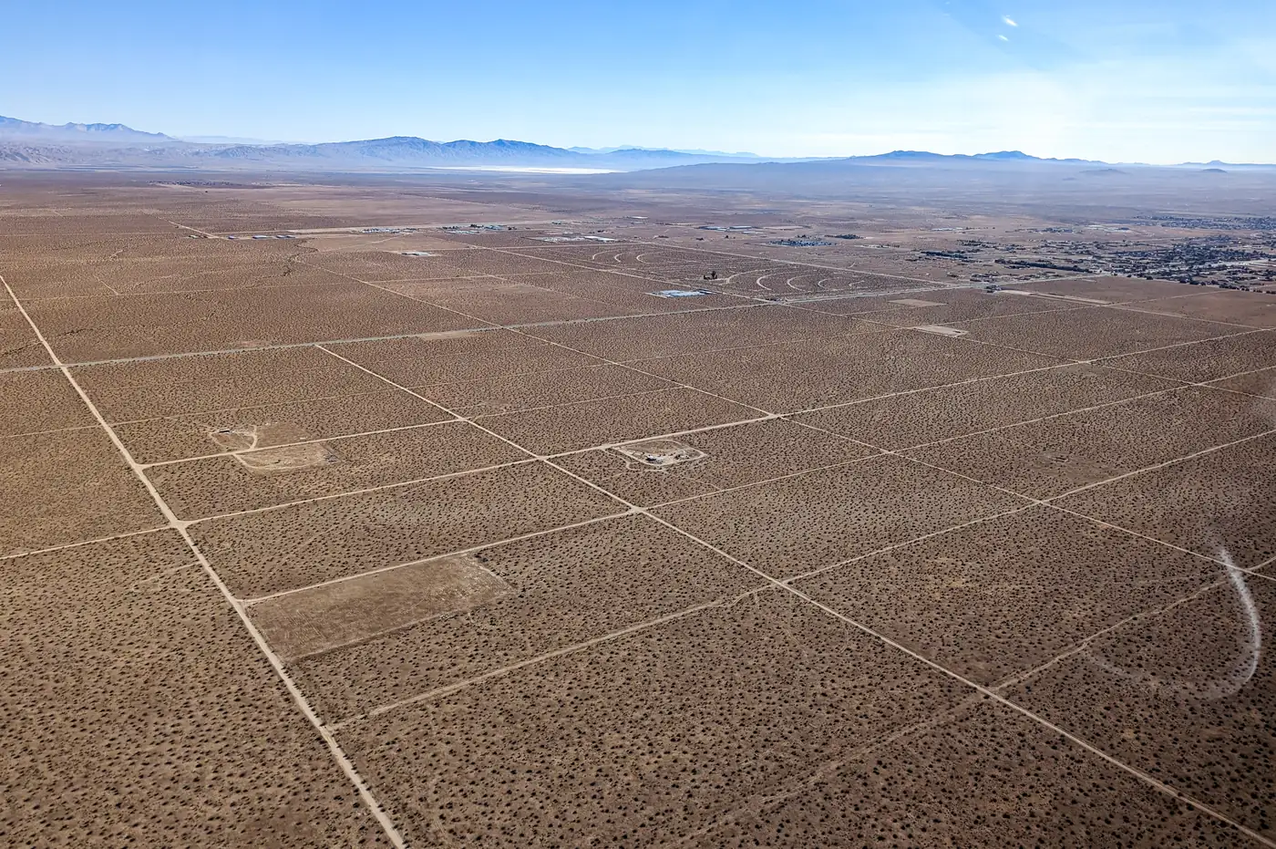 Aerial photograph of California City's empty grid: straight graded dirt streets cross flat creosote desert in a pattern of large blocks, with only two or three small clearings on them, and the built-up town a thin band on the far right beneath distant mountains.