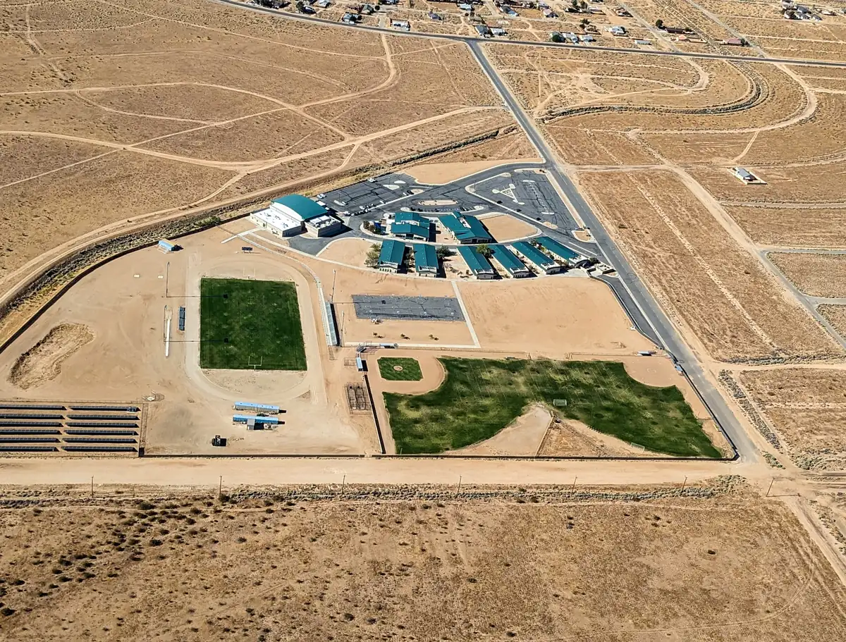 Aerial photograph of California City High School: green-roofed classroom buildings and a gym around a car park, a grass football field inside a running track, a baseball field and rows of solar panels, surrounded by bare desert crossed by dirt roads, with houses along the top edge.
