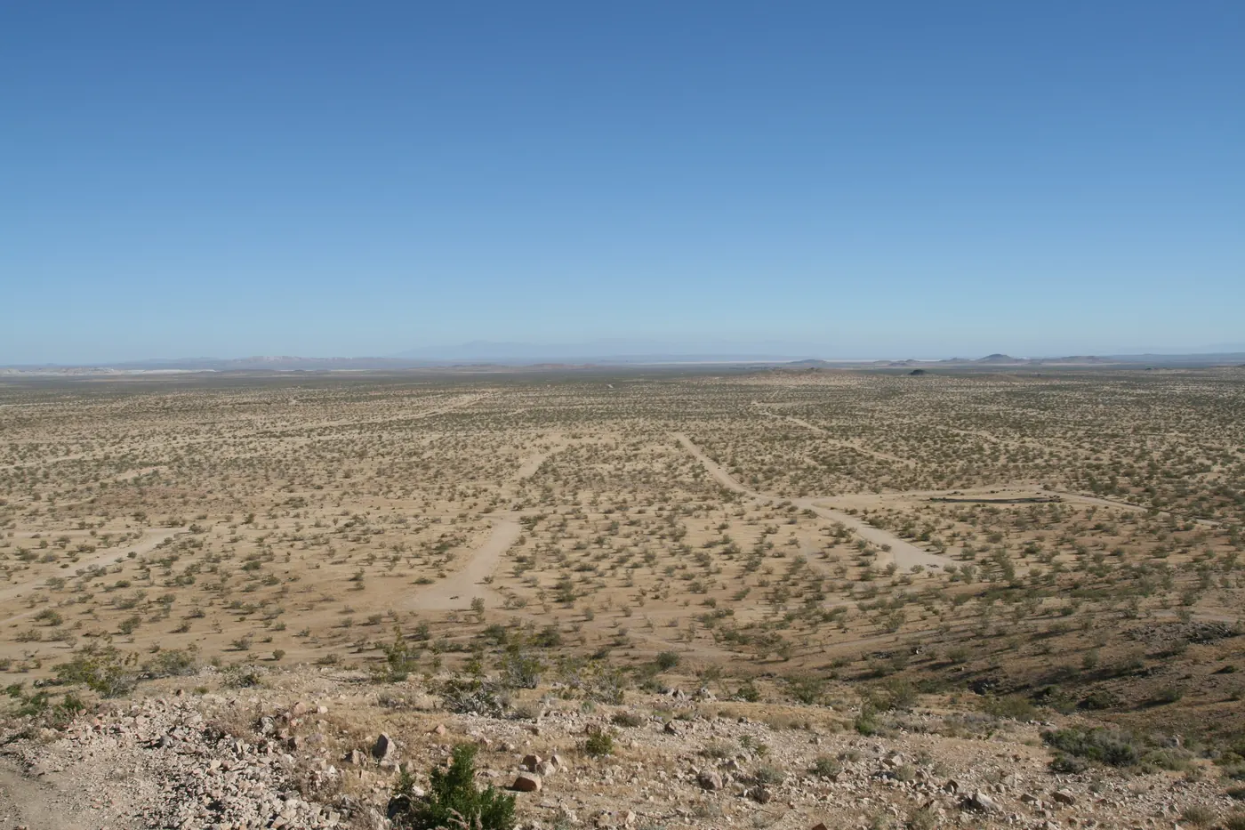 Looking down from a rocky hillside over flat desert scrub stretching to a far horizon, crossed by the pale lines of dirt streets, with no buildings anywhere.