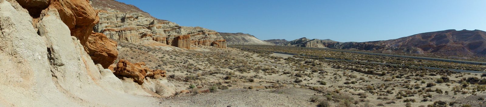 Wide panorama of pale and orange eroded rock close by on the left, opening onto a sandy scrub-covered flat, a highway and a line of cliffs and hills running off to the right.