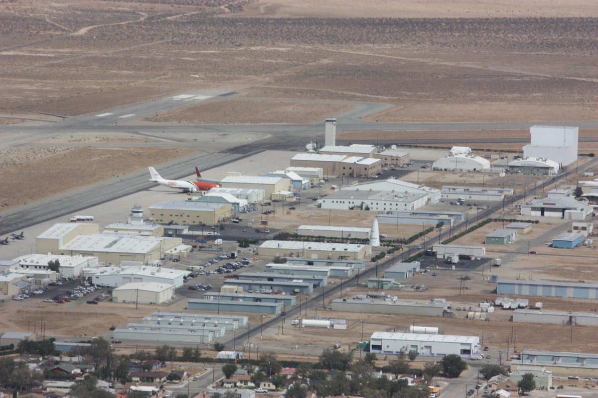Aerial photograph of an airfield on a desert plain: rows of hangars and low industrial buildings, parked jet airliners, runways and a control tower, with the edge of a town in the foreground.