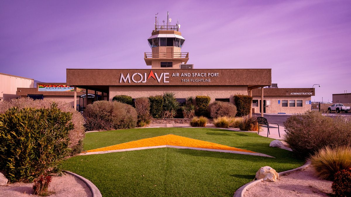 A low brown building lettered Mojave Air and Space Port, 1434 Flightline, with a control tower rising behind it, a Voyager Restaurant sign on the left and an Administration wing on the right, behind a lawn and shrubs under a purple-tinted sky.