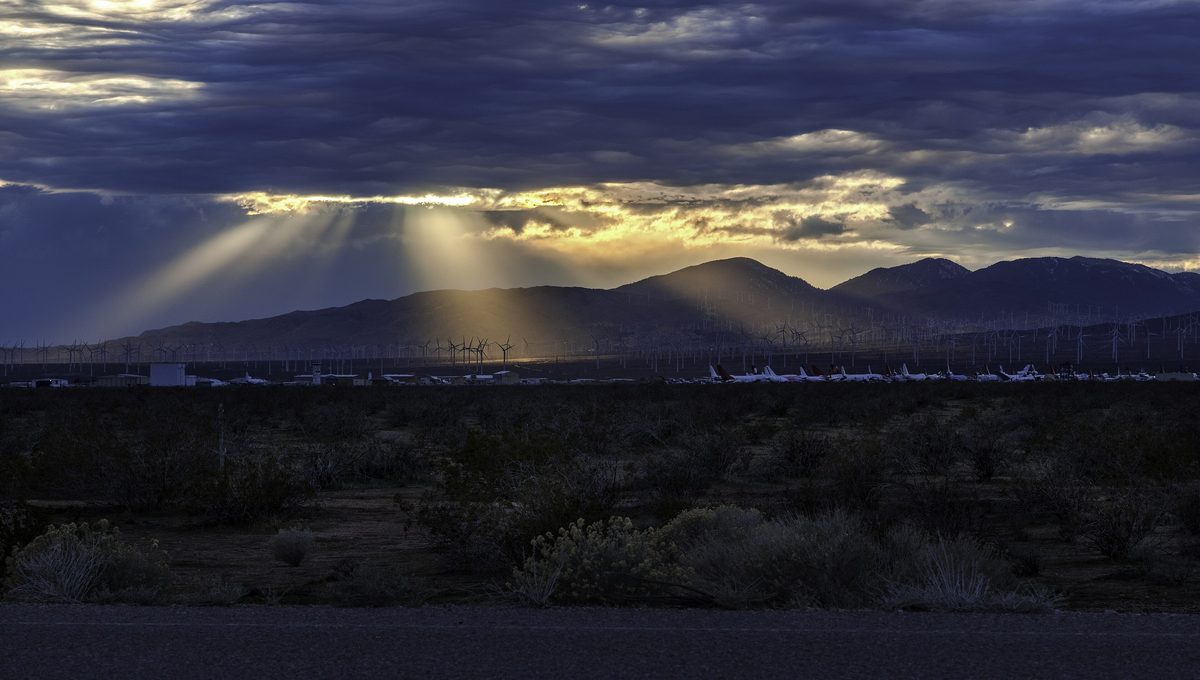 Rows of parked airliners seen in silhouette across a dark desert plain, under storm clouds broken by shafts of sunlight over the mountains beyond.