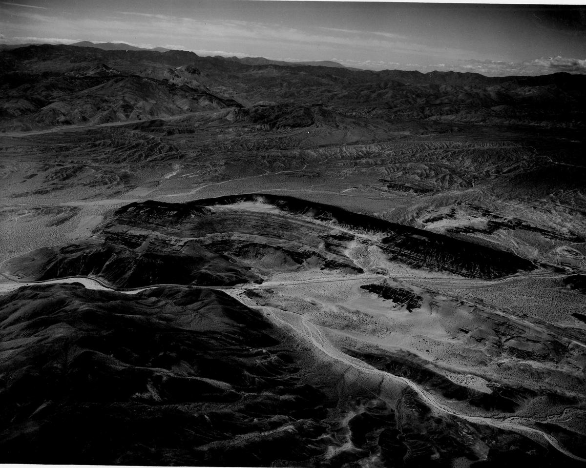 Black-and-white aerial photograph of eroded desert ridges and canyons, with a road winding through them and mountains on the horizon.