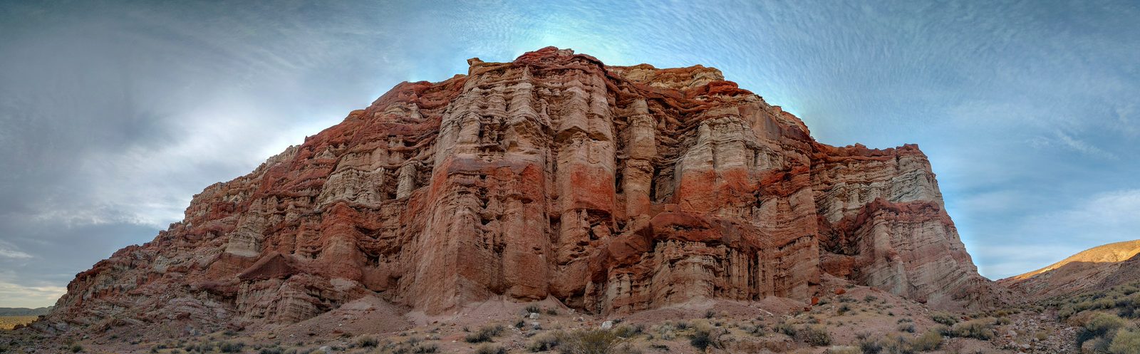 Panorama of a tall butte of fluted cliffs banded in rust red and pale cream, rising from scrubby desert under a streaked sky.