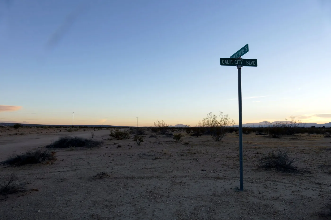 A green street sign reading Calif City Blvd and Country Club Dr on a pole in bare desert scrub at dusk, with power poles and distant mountains on the horizon.