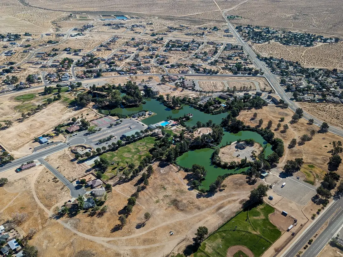 Aerial photograph of Central Park in California City: a winding green lake with a tree-covered island, a swimming pool, tennis courts, a car park, a baseball field and lawns, surrounded by houses on curving streets and open desert beyond.