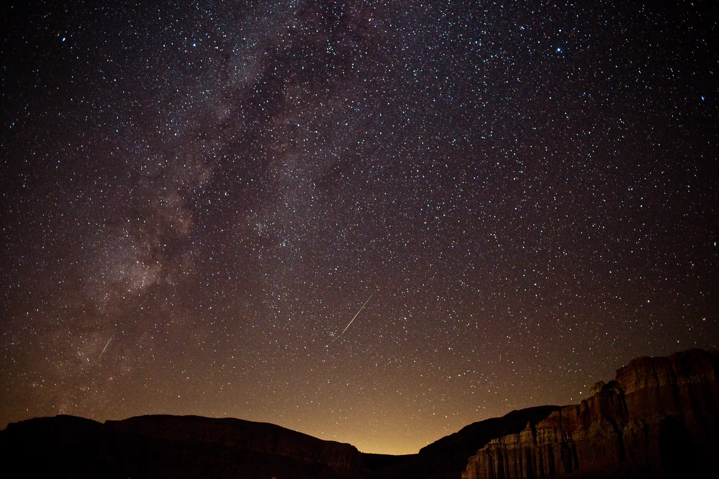 A night sky packed with stars over the dark silhouette of eroded cliffs, with the band of the Milky Way rising on the left and the bright streaks of two meteors.