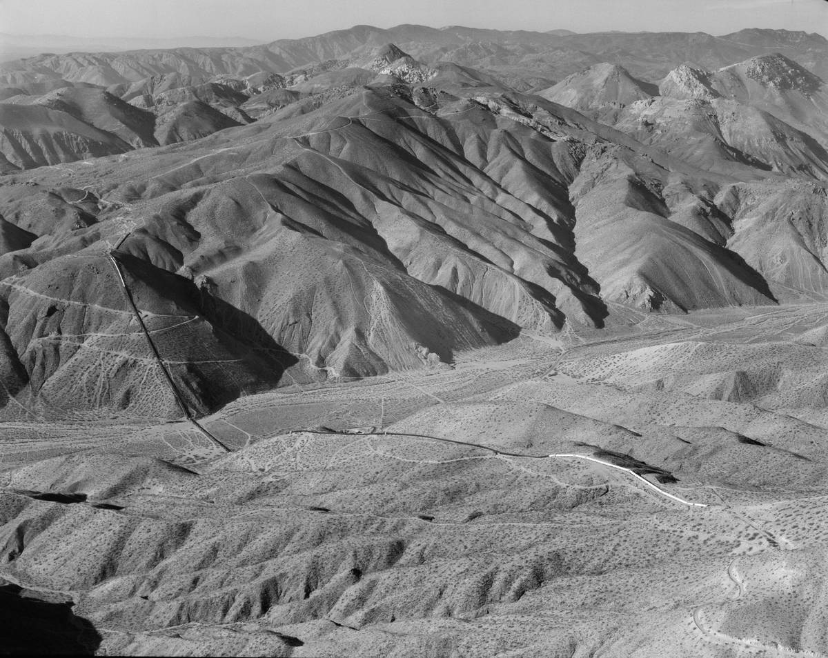 Black-and-white aerial photograph of steep, furrowed desert mountains, with a dark straight line running down one slope and across the canyon floor beside a winding road.
