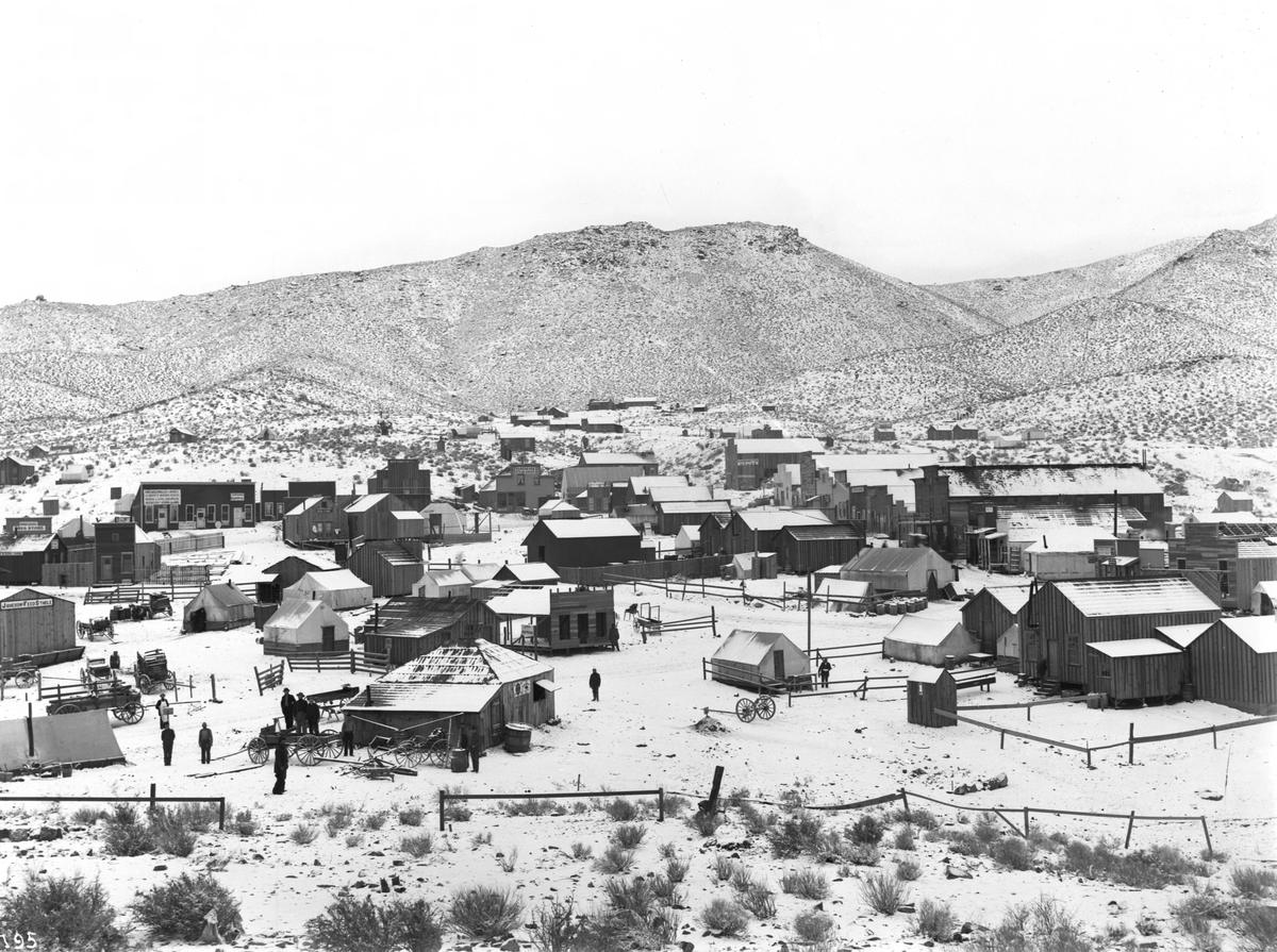 Black-and-white photograph of a small mining town of wooden buildings and tents on snowy ground, with a few people walking between them and bare hills behind.